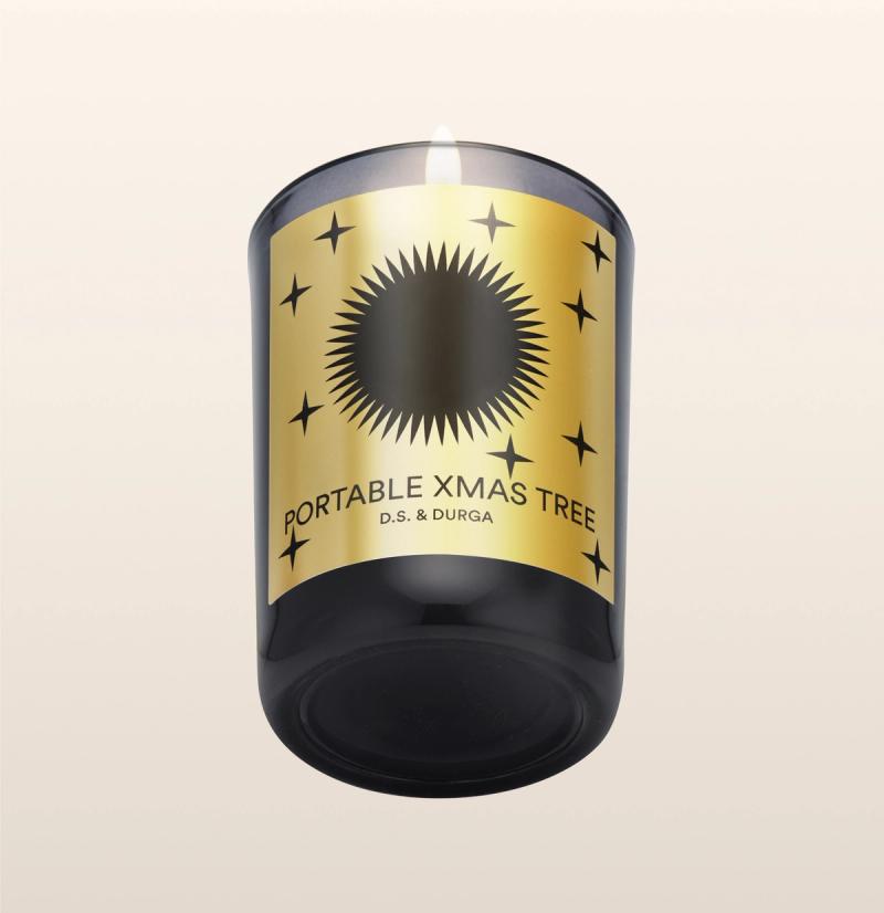 A lit candle in a dark glass holder with a gold label. The label features a black sunburst design and small stars, with the text "Portable Xmas Tree" and "D.S. & Durga" on a beige gradient background.