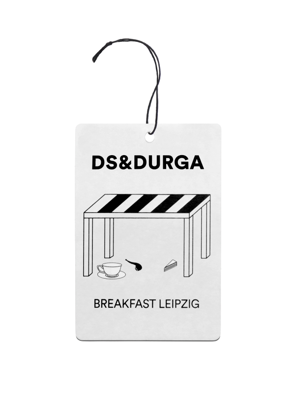 A hanging auto fragrance with the inscription"d.s. & durga" on the upper portion and "breakfast leipzig" below featuring stylish monochrome design with a table, cup, and spoon illustration