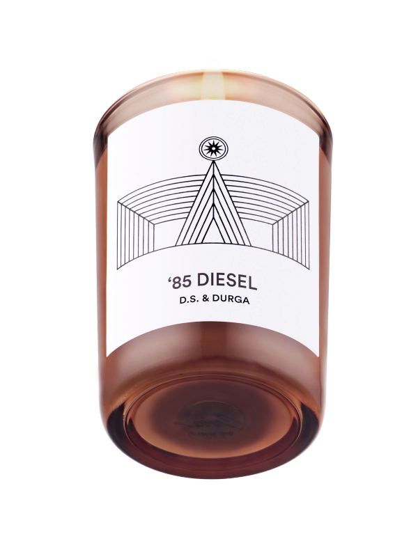 A lit scented candle with a sleek design, featuring the label '85 diesel d.s. & durga', which suggests a unique fragrance inspired by the named theme.