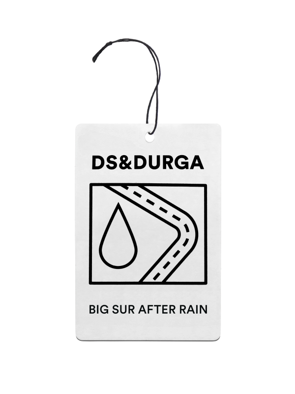 A hanging auto fragrance with the inscription "ds &amp; durga" and "big sur after rain" on it, featuring a stylized road and raindrop graphic.