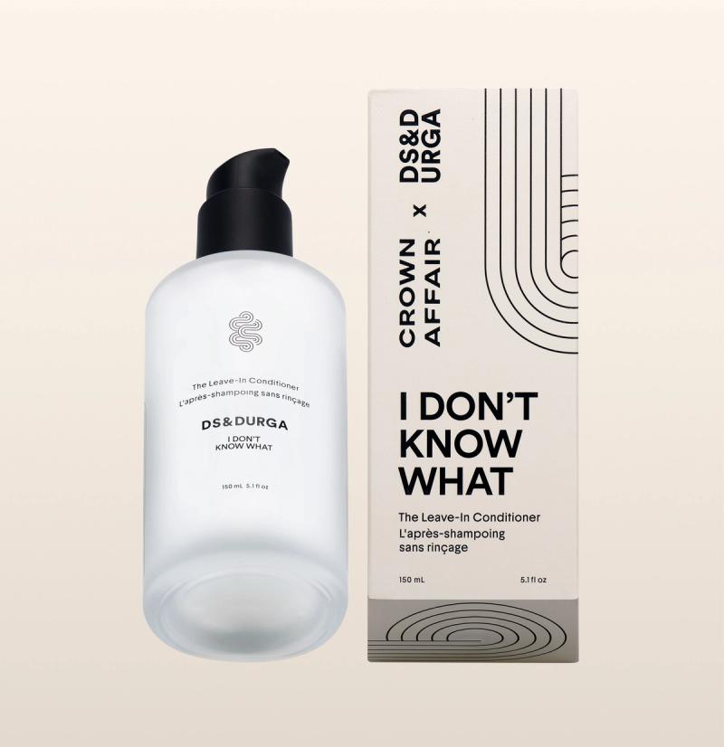 A bottle of DS & Durga x Crown Affair "I DON'T KNOW WHAT" leave-in conditioner stands next to its beige, black-striped box on a light background. The bottle has a black pump and minimalist label.