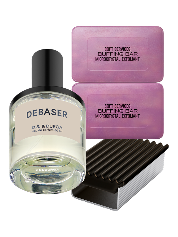 An arrangement of D.S. & Durga Debaser eau de parfum bottle and two Soft Services buffing bar microcrystal exfoliants. The perfume bottle is in the foreground, while the two purple buffing bars and their dark purple ridged holder are in the background.