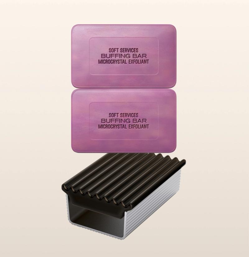 Two rectangular pink bars labeled "Soft Services Buffing Bar Microcrystal Exfoliant" are positioned above a dark purple and white ribbed soap holder. The bars are stacked one on the other, with the soap holder placed beneath.