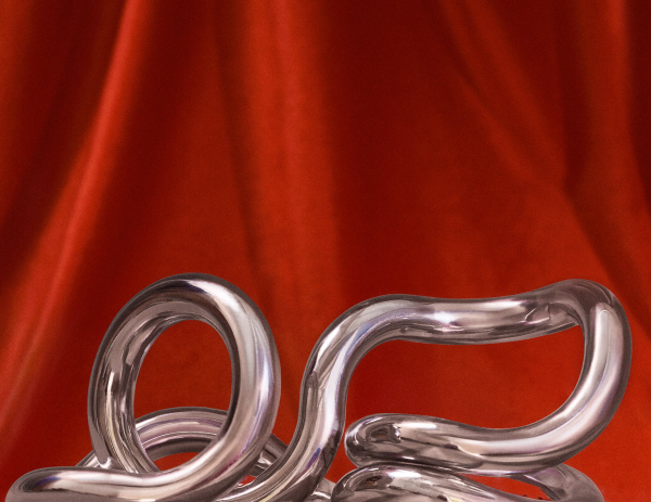A shiny, abstract silver sculpture with looping curves is displayed against a backdrop of rich red fabric with soft folds.