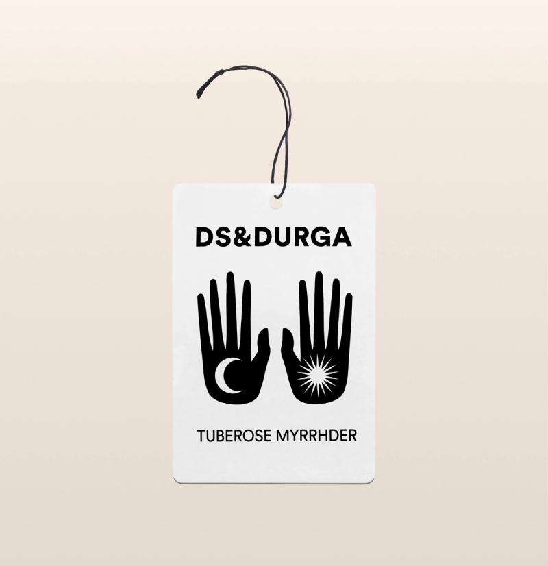 A hanging auto fragrance bold typography showcasing the brand "ds & durga" and the product name "tuberose myrrhder". the label features two stylized, symmetrical icons of an open hand, one with an eye in the palm and the other with a radiating star or burst.