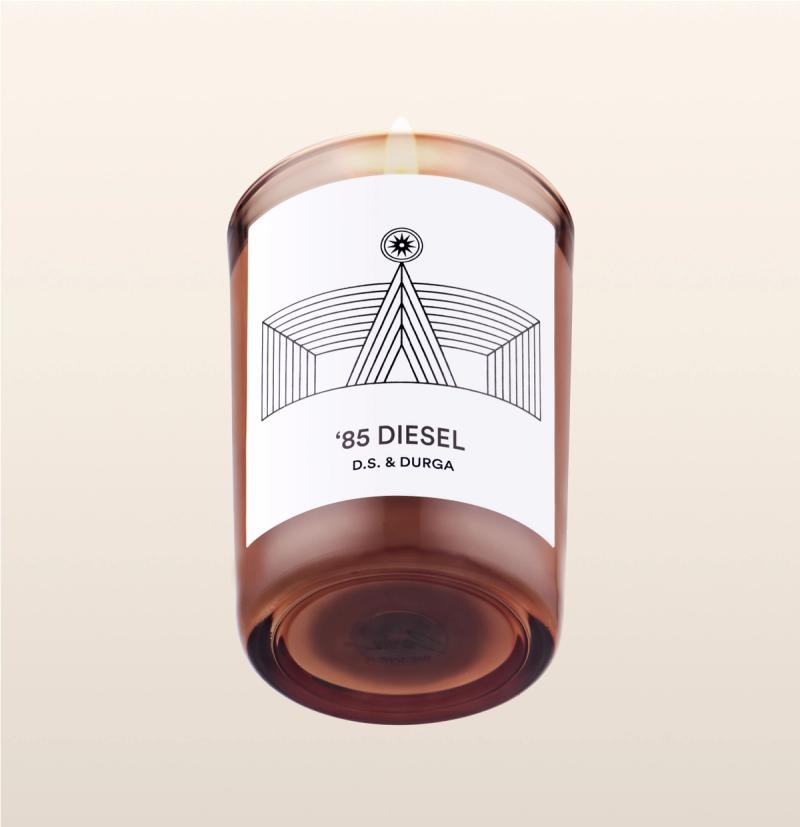 A lit scented candle with a sleek design, featuring the label '85 diesel d.s. & durga', which suggests a unique fragrance inspired by the named theme.