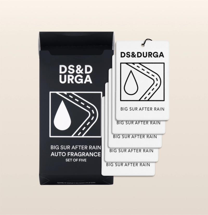 A black box labeled "DS & DURGA" containing five white air fresheners with the same design of a raindrop on a winding road and labeled "BIG SUR AFTER RAIN AUTO FRAGRANCE SET OF FIVE." The box and air fresheners are set against a beige background.