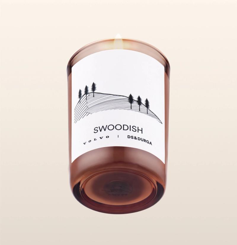 A tan glass candle with a white label showing a hill with trees sketched on it. The label reads "SWOODISH Volvo | DS&DURGA." The candle is lit, with a small flame visible at the top. The background is a light beige color.