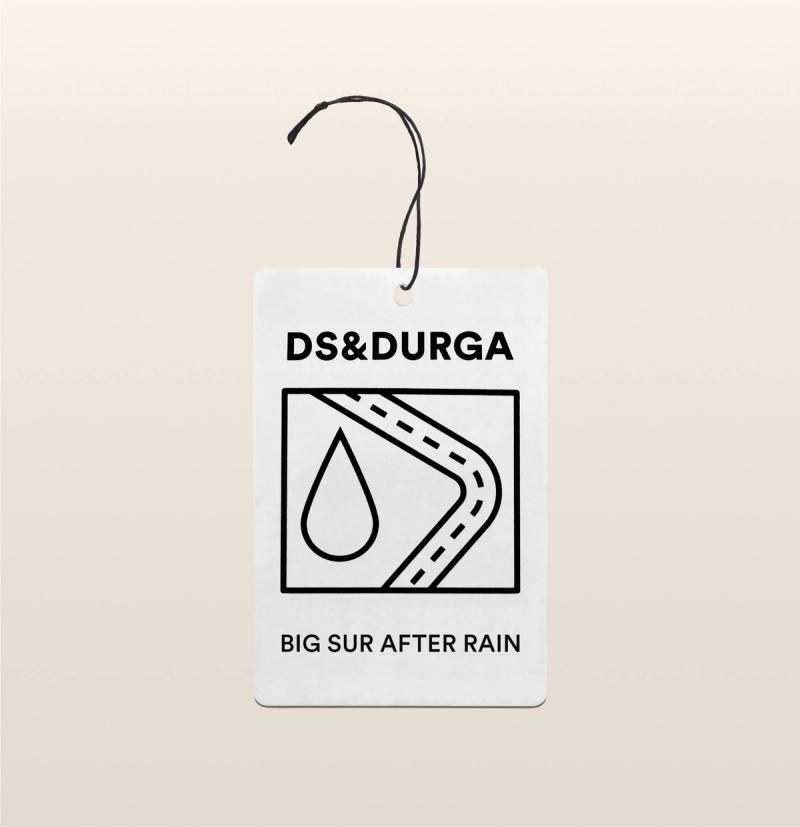 A hanging auto fragrance with the inscription "ds &amp; durga" and "big sur after rain" on it, featuring a stylized road and raindrop graphic.