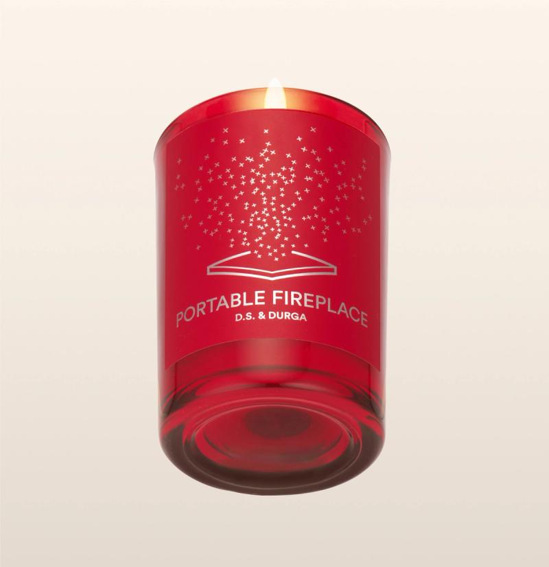 A red glass candle labeled "PORTABLE FIREPLACE D.S. &amp; DURGA," with a flame burning and a pattern of small stars above an open book design on the front. The background is plain and light-colored.
