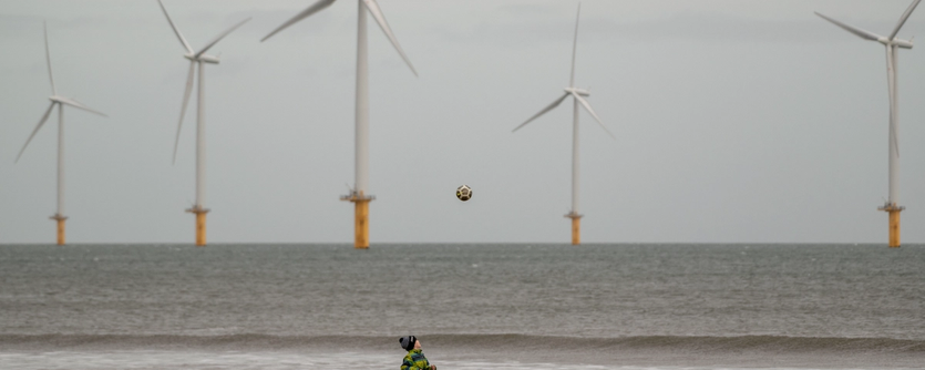 A boy plays football on a beach with an offshore wind farm in the background