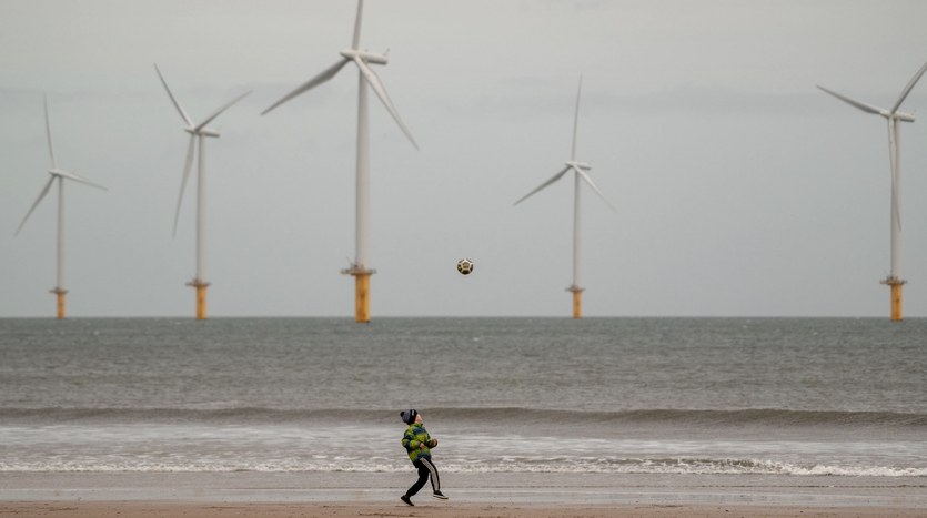 A boy plays football on a beach with an offshore wind farm in the background