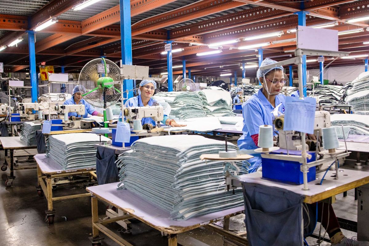 Workers sew fabric at machines amid stacks of folded textiles in a brightly lit garment factory.