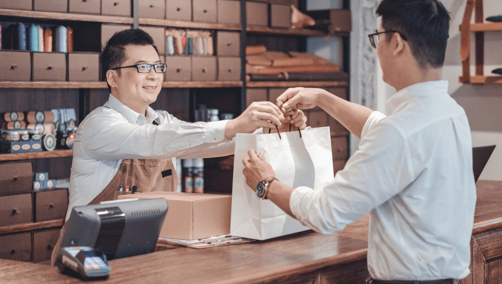 Smiling shop clerk hands a white shopping bag to a customer across the counter