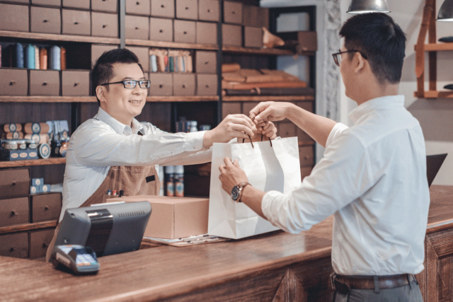Smiling shop clerk hands a white shopping bag to a customer across the counter