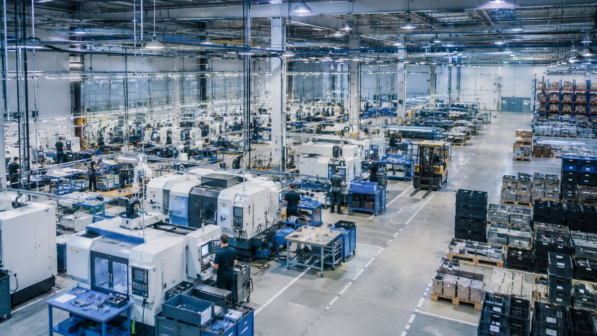 Large industrial factory floor with workers operating CNC machines and rows of pallets and storage racks