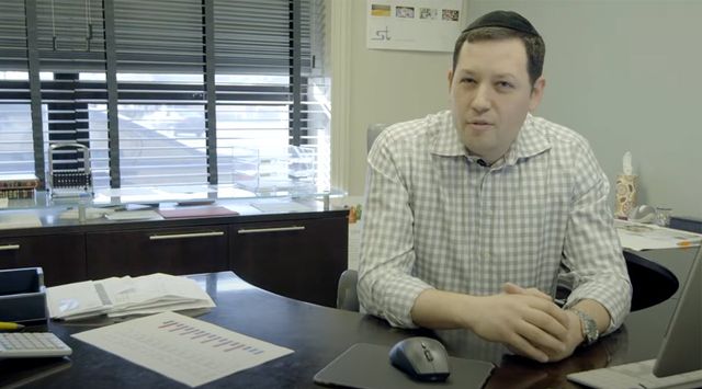 Man in checkered shirt sits at office desk with papers, charts, and a computer mouse in front of him.