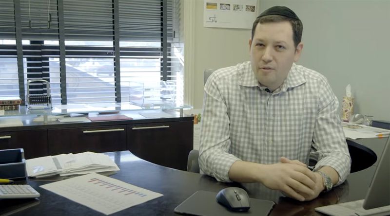 Man in checkered shirt sits at office desk with papers, charts, and a computer mouse in front of him.
