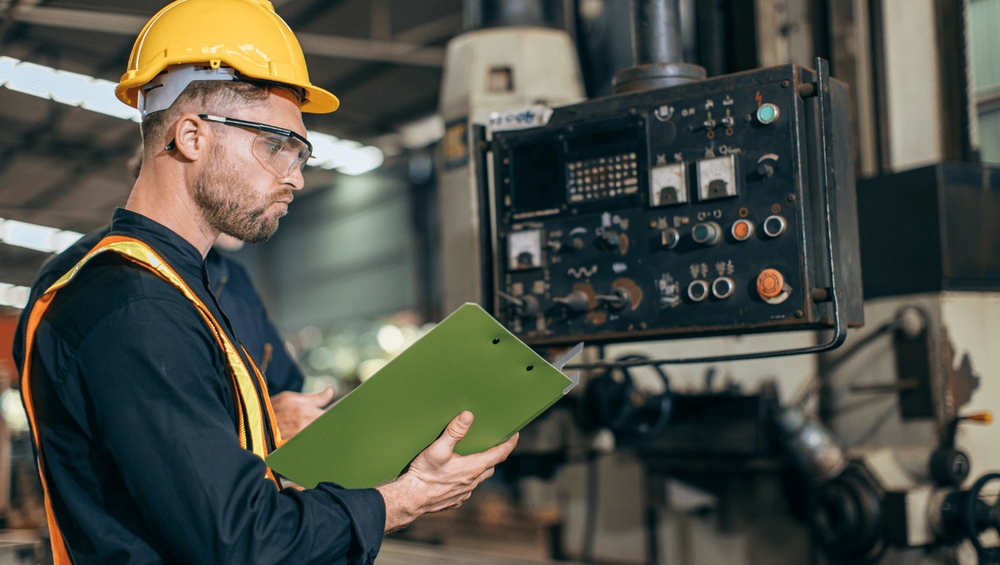 Factory worker in hard hat and vest inspects control panel while holding a green clipboard