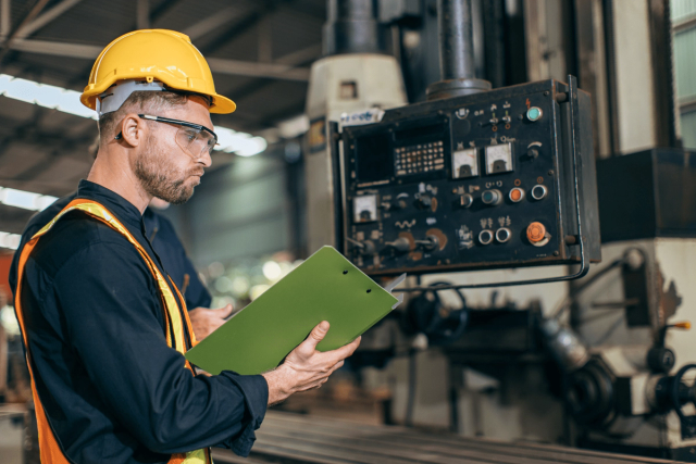 Factory worker in hard hat and vest inspects control panel while holding a green clipboard