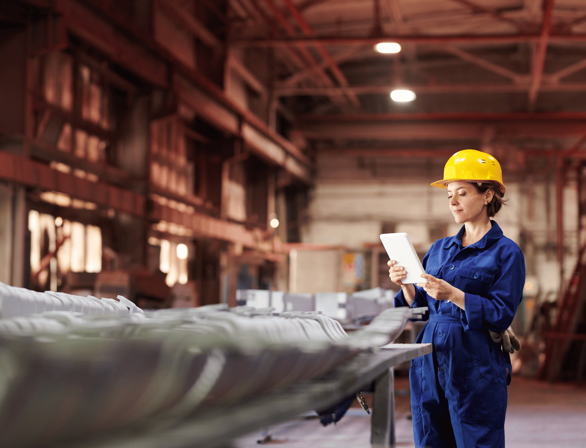 Factory worker in blue coveralls and yellow hard hat using a tablet beside a production line