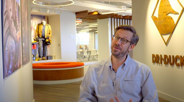Man in glasses speaks in a modern office lobby with DRI DUCK logo and clothing display behind him