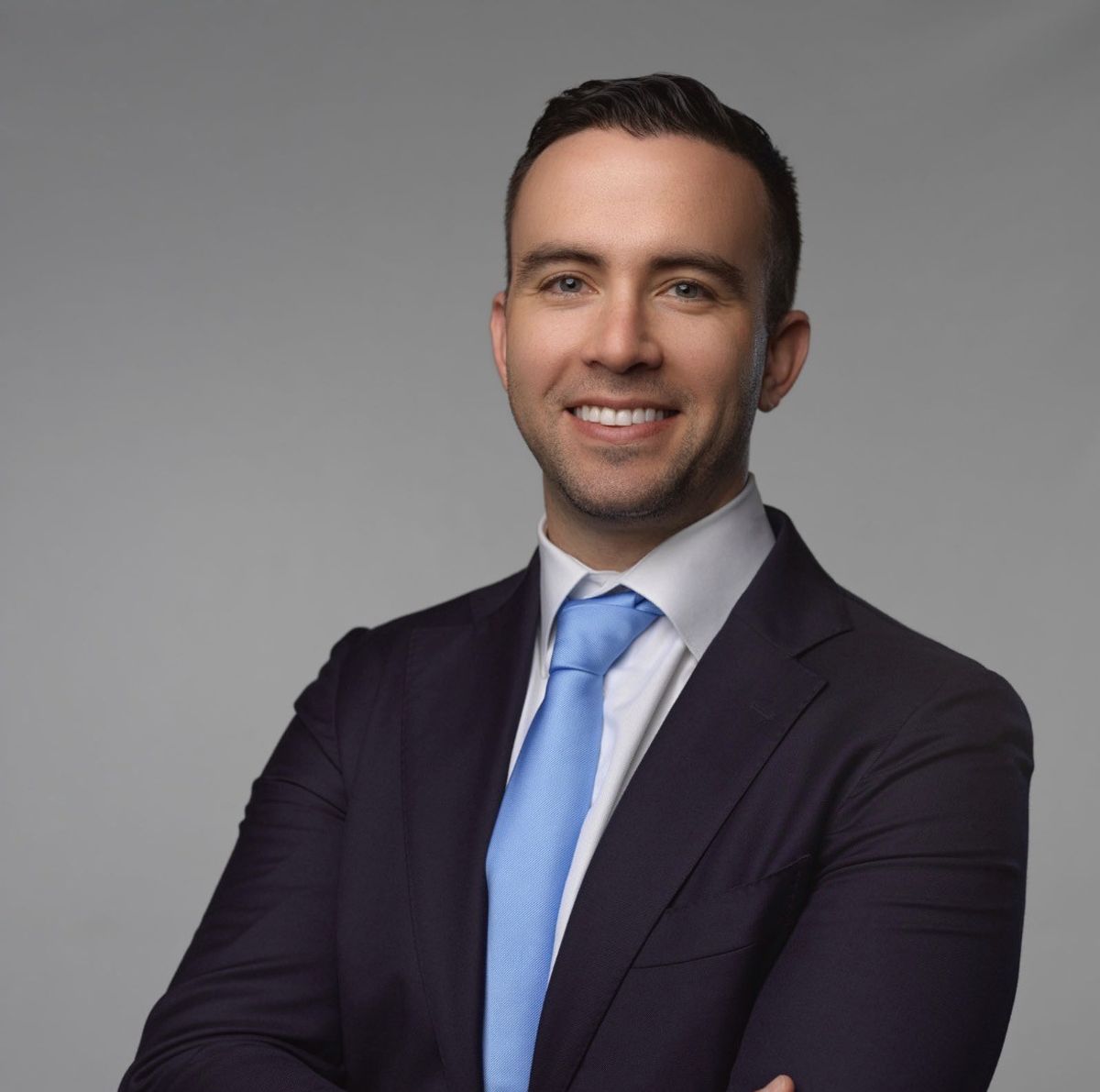Smiling man in a dark suit and light blue tie posing against a plain gray background