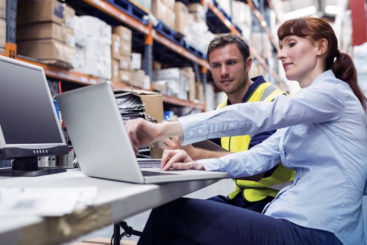 Two warehouse workers at a desk using laptops and a monitor, with shelves of boxes in the background
