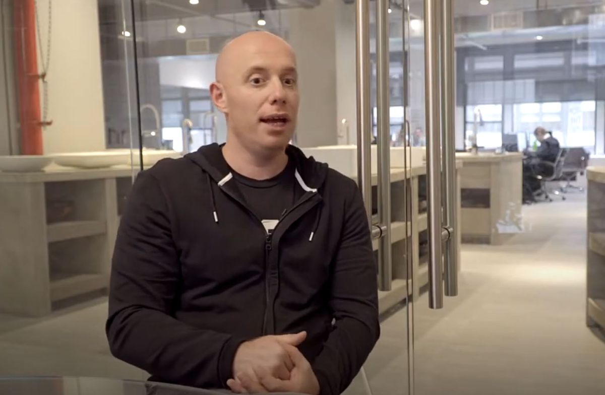 Bald man in a black hoodie speaks while seated in a modern glass-walled office.