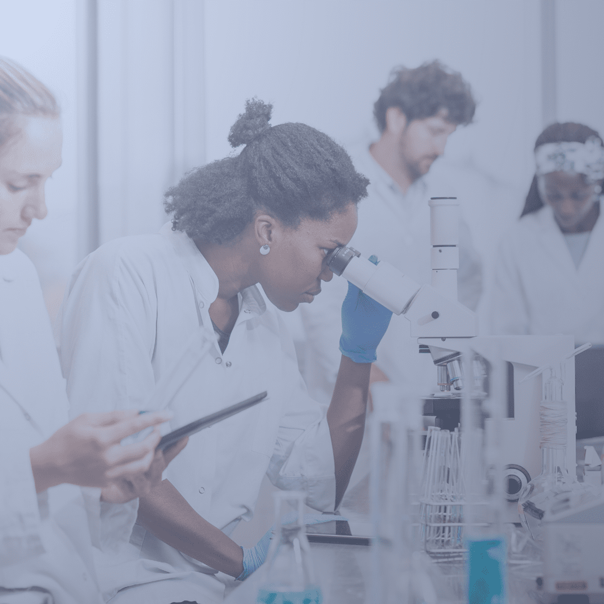 Scientists in lab coats work at a bench while a woman examines a sample through a microscope