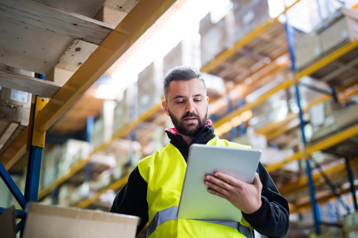 Warehouse worker in a high-visibility vest checking inventory on a tablet among tall storage shelves