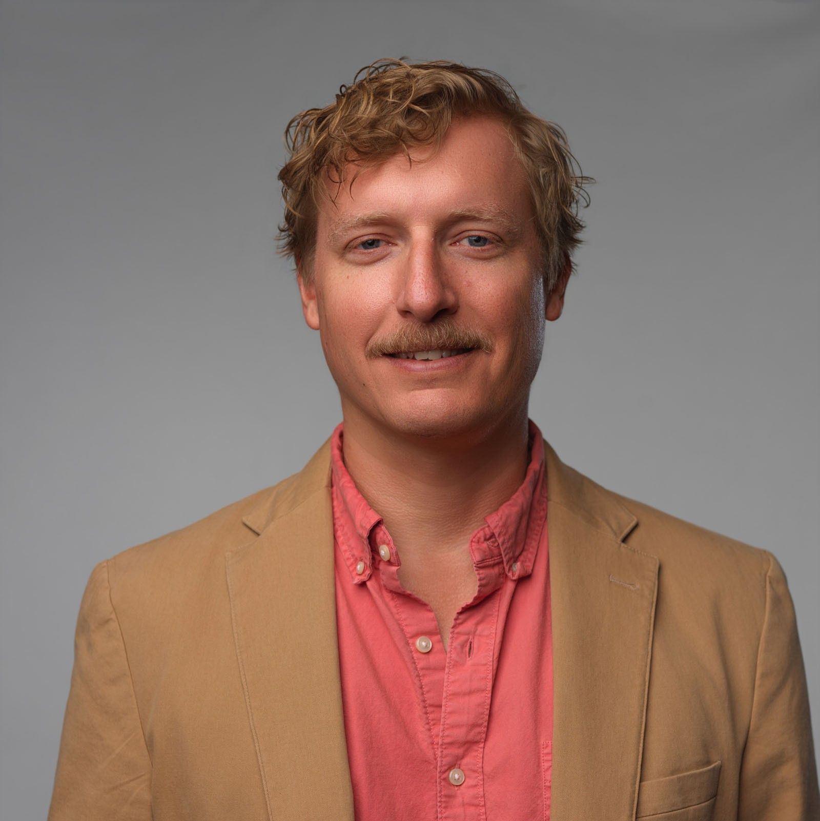 Man with wavy blond hair and mustache wearing tan blazer and coral shirt against plain gray background