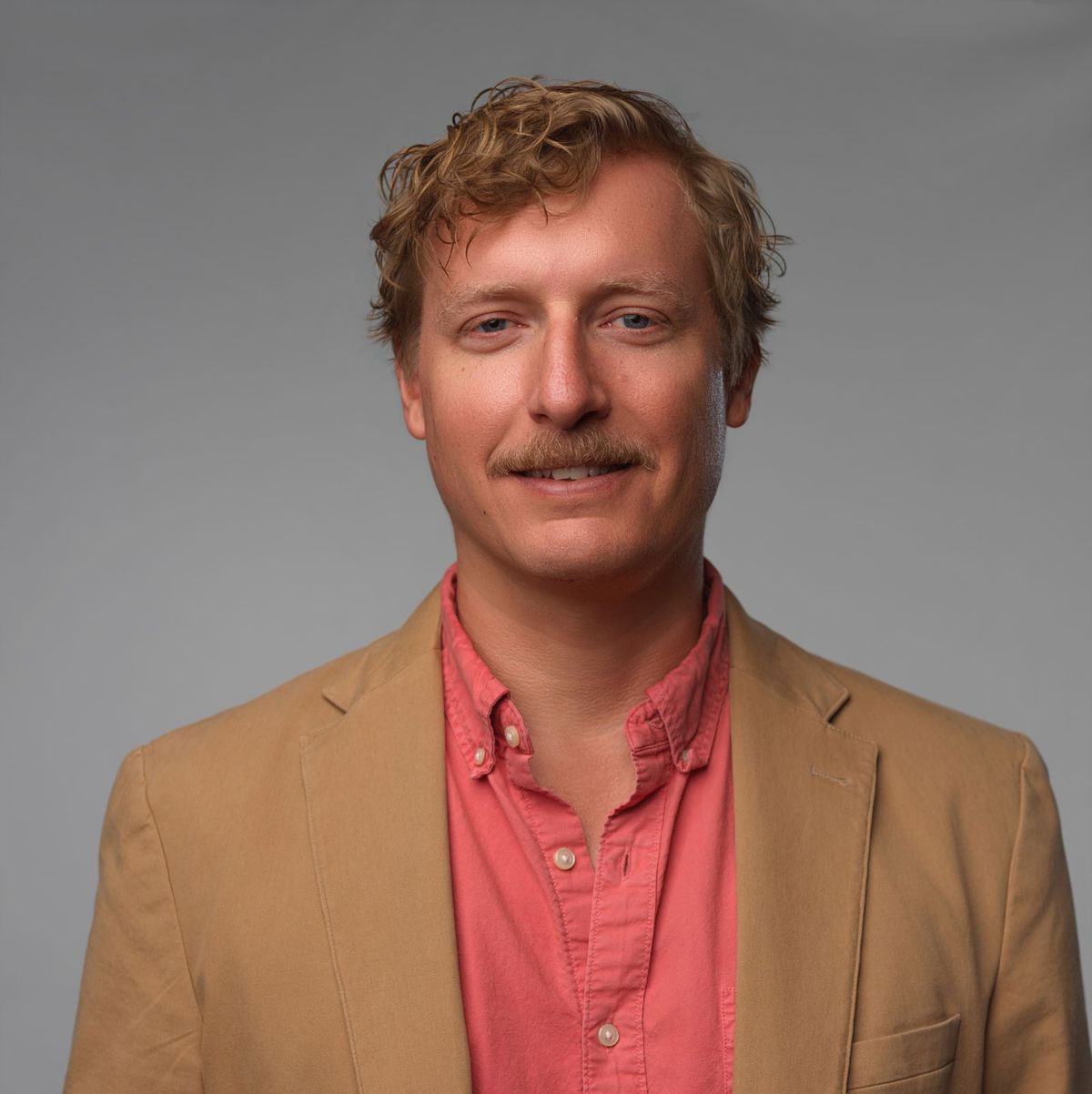 Man with wavy blond hair and mustache wearing tan blazer and coral shirt against plain gray background