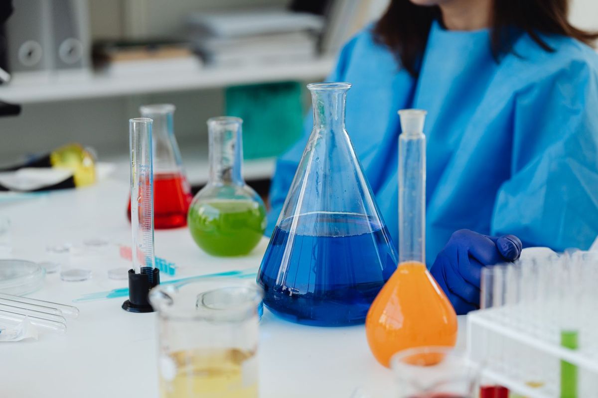 Scientist in blue protective gear works among laboratory glassware filled with colorful liquids.