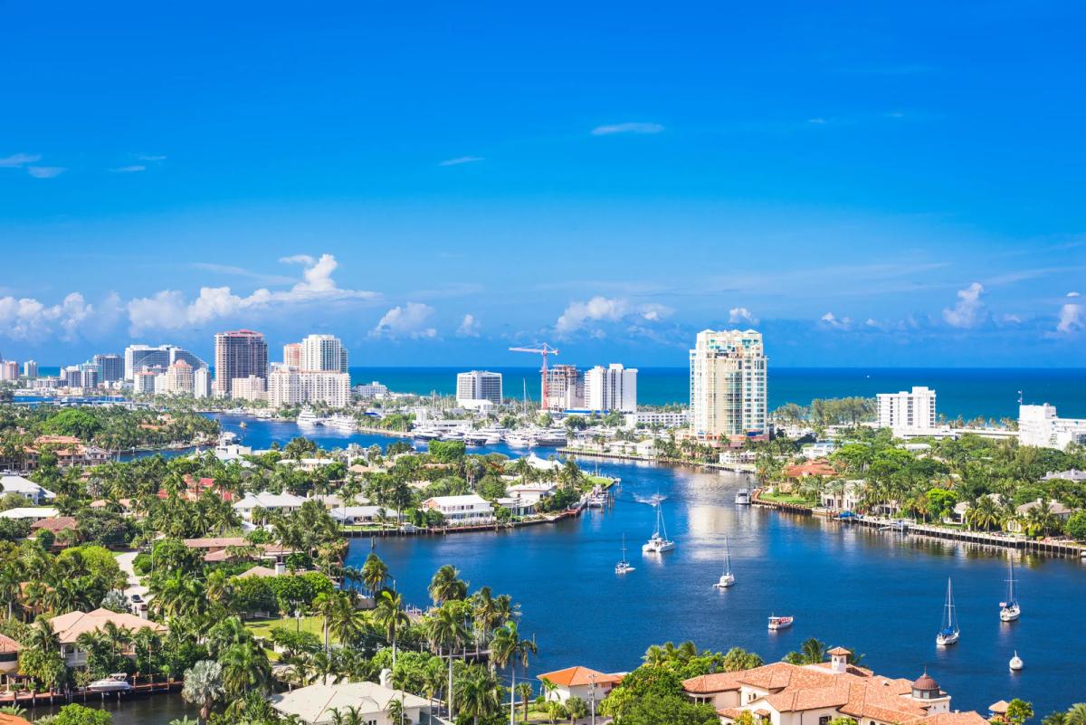 Coastal city skyline with canals, yachts, and sailboats surrounded by palm trees under a bright blue sky
