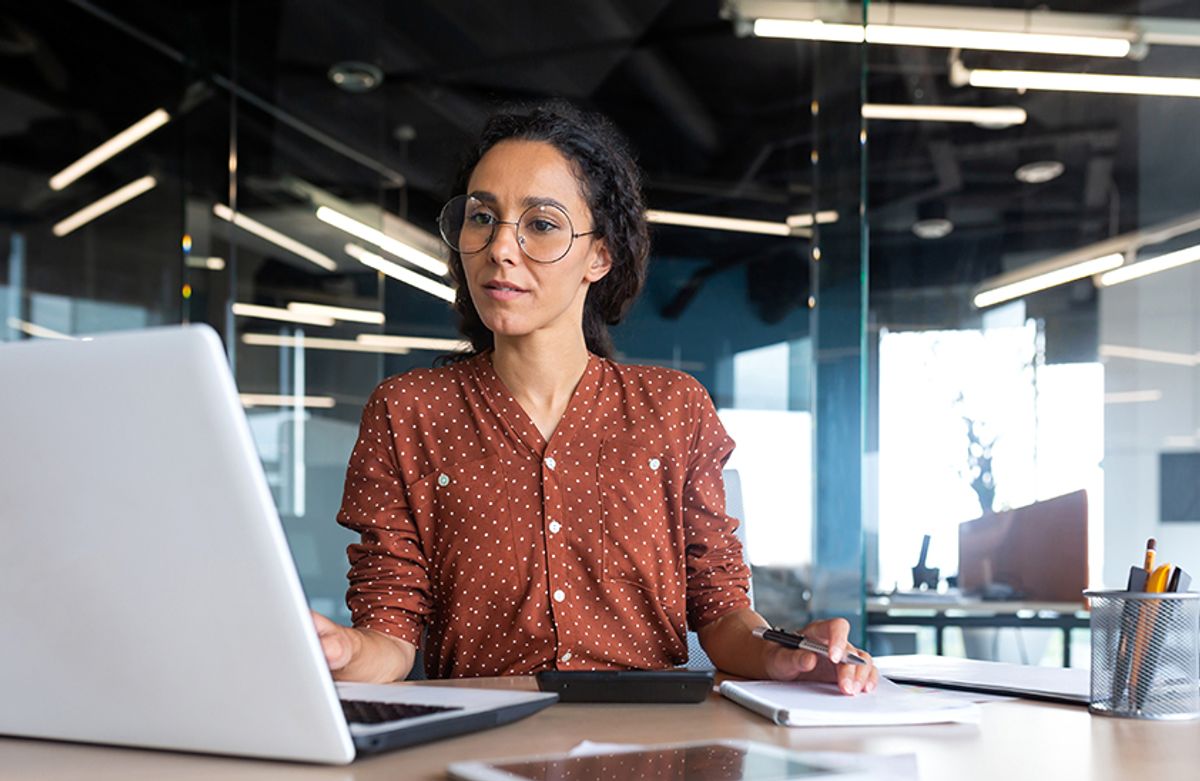 Woman in glasses working at a laptop in a modern office with glass walls and overhead lights