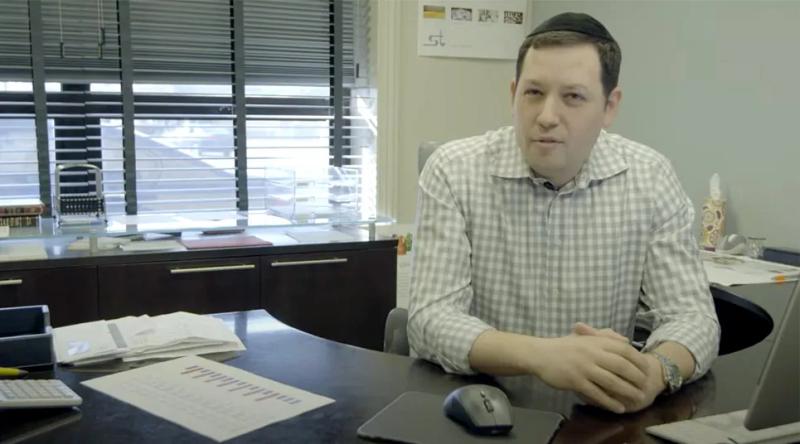 Man in checkered shirt sits at office desk with papers, computer mouse, and window blinds behind him