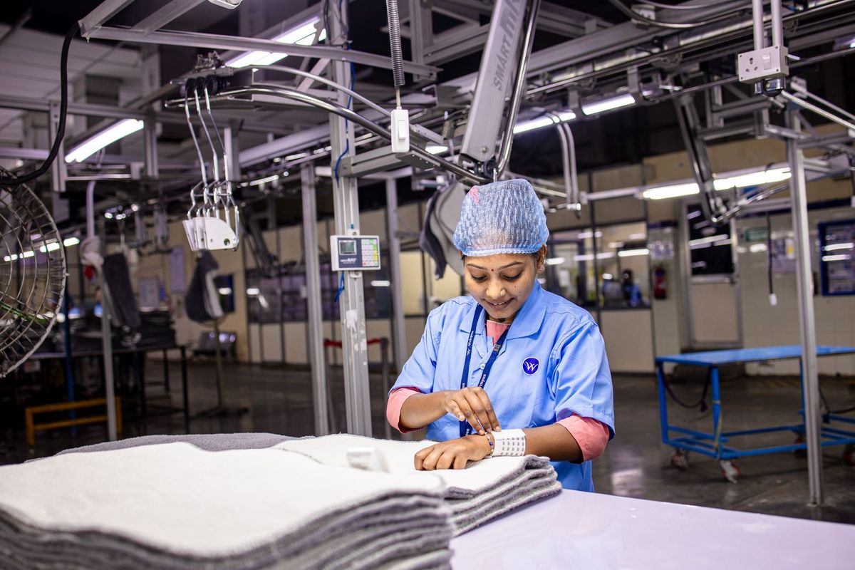 Factory worker in a blue uniform and hairnet inspects folded fabric at an industrial workstation.