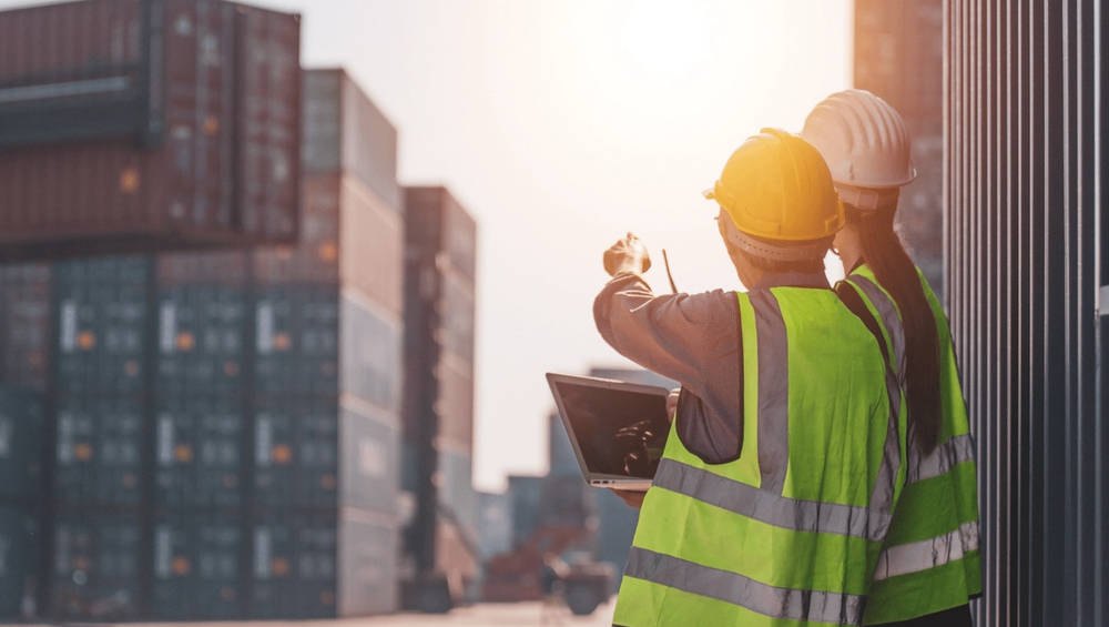 Two workers in safety vests and helmets inspect shipping containers at a sunlit freight yard