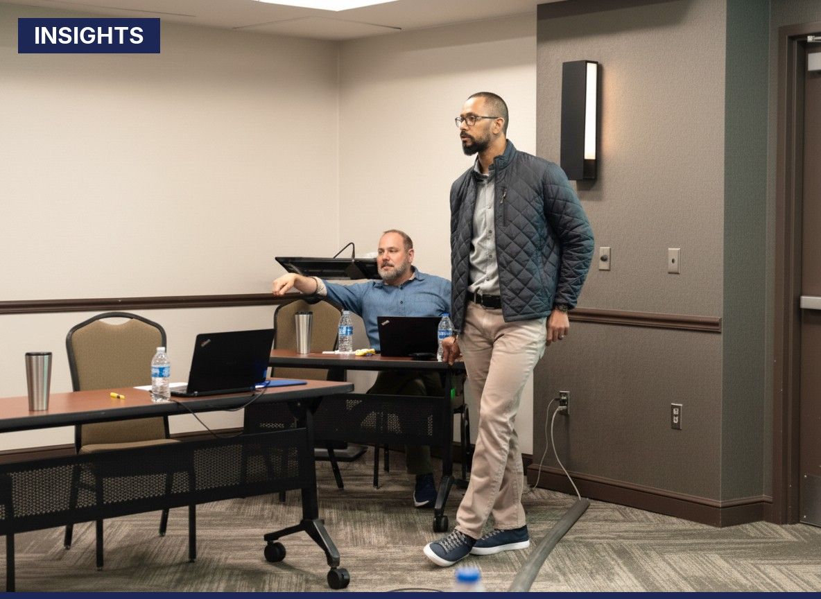 Man standing and speaking in a conference room while another man sits at a table with laptops and water bottles.