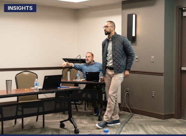 Man standing and speaking in a conference room while another man sits at a table with laptops and water bottles.