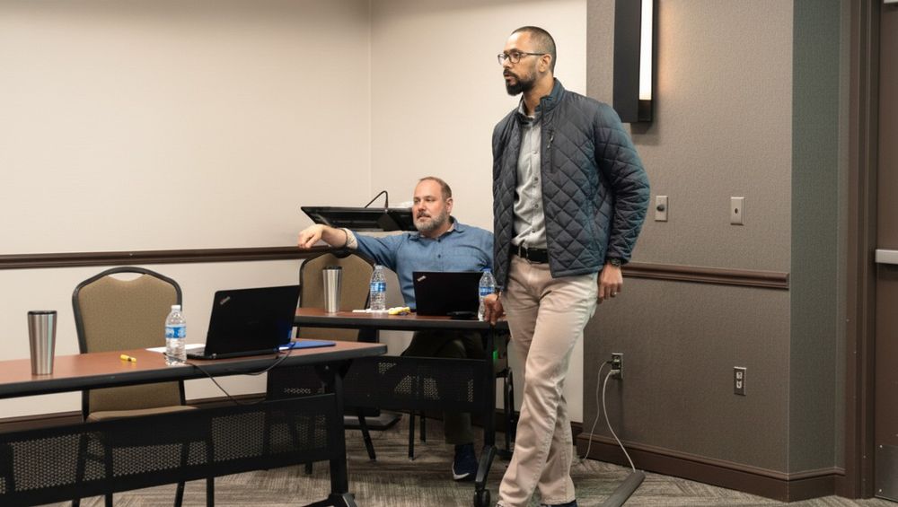 Man standing and speaking in a conference room while another man sits at a table with laptops and water bottles.
