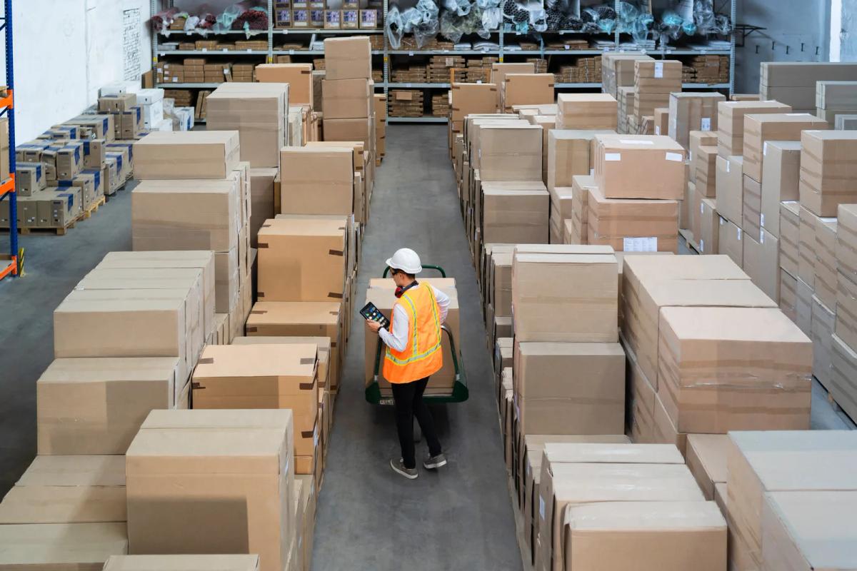 Warehouse worker in safety vest and hard hat checks a tablet while moving boxes on a cart between tall stacks