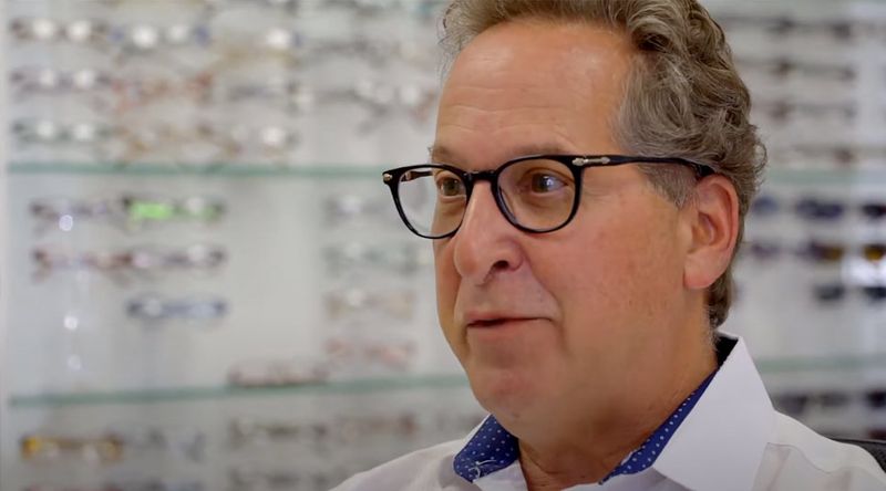 Middle-aged man wearing black glasses in an eyewear shop with display shelves behind him