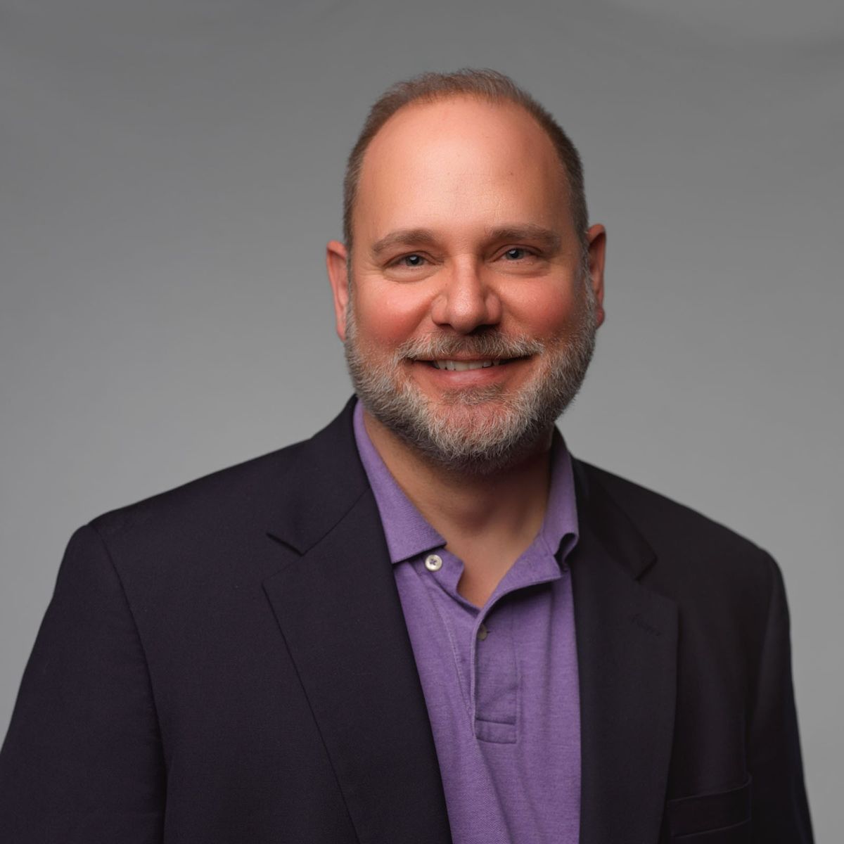 Smiling man with short hair and beard wearing a purple polo and dark blazer against a plain gray background