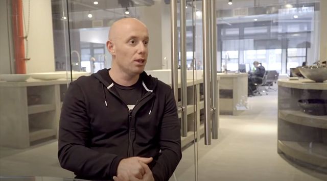 Bald man in a black hoodie speaking while seated in a modern glass-walled office