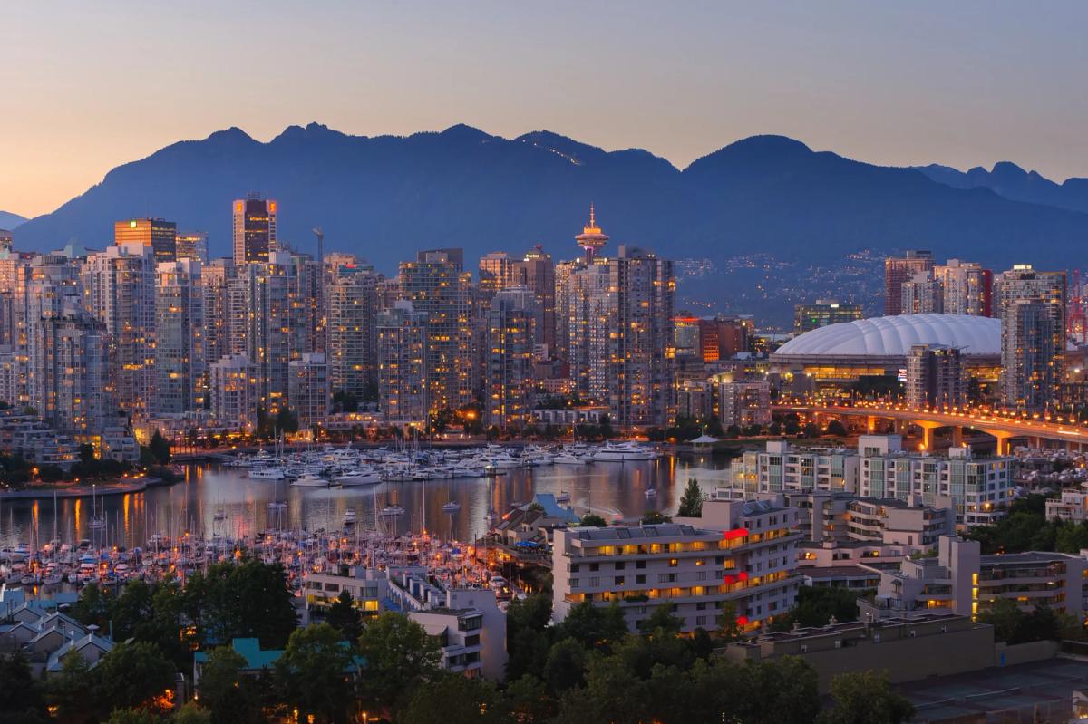 City skyline at dusk with lit high-rises, marina boats, and mountains silhouetted in the background