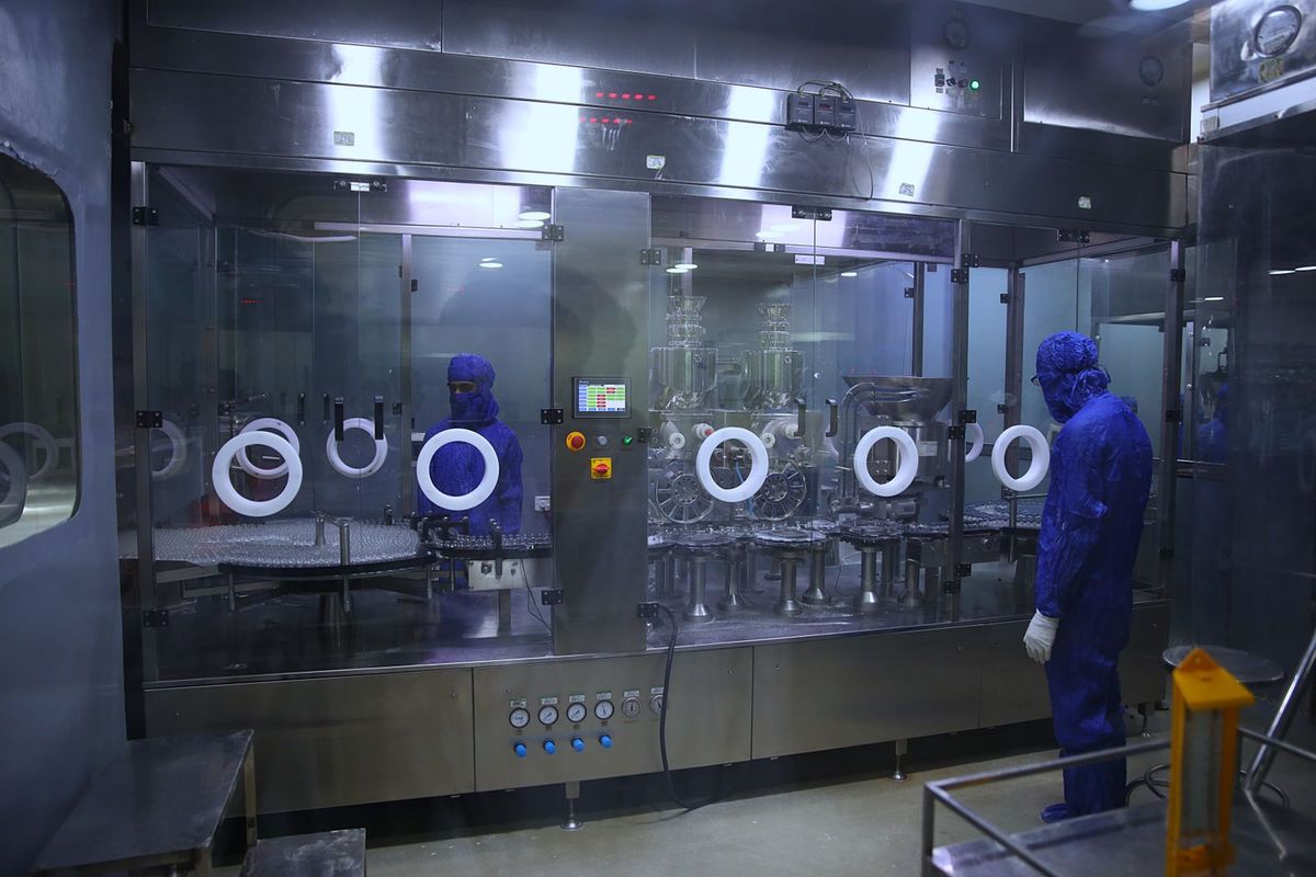 Workers in blue protective suits monitor an enclosed pharmaceutical bottling machine.