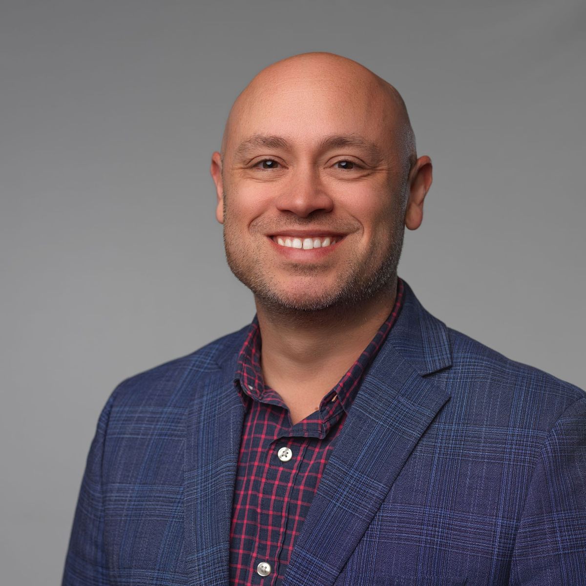 Smiling bald man in a blue plaid blazer and red checkered shirt against a gray background