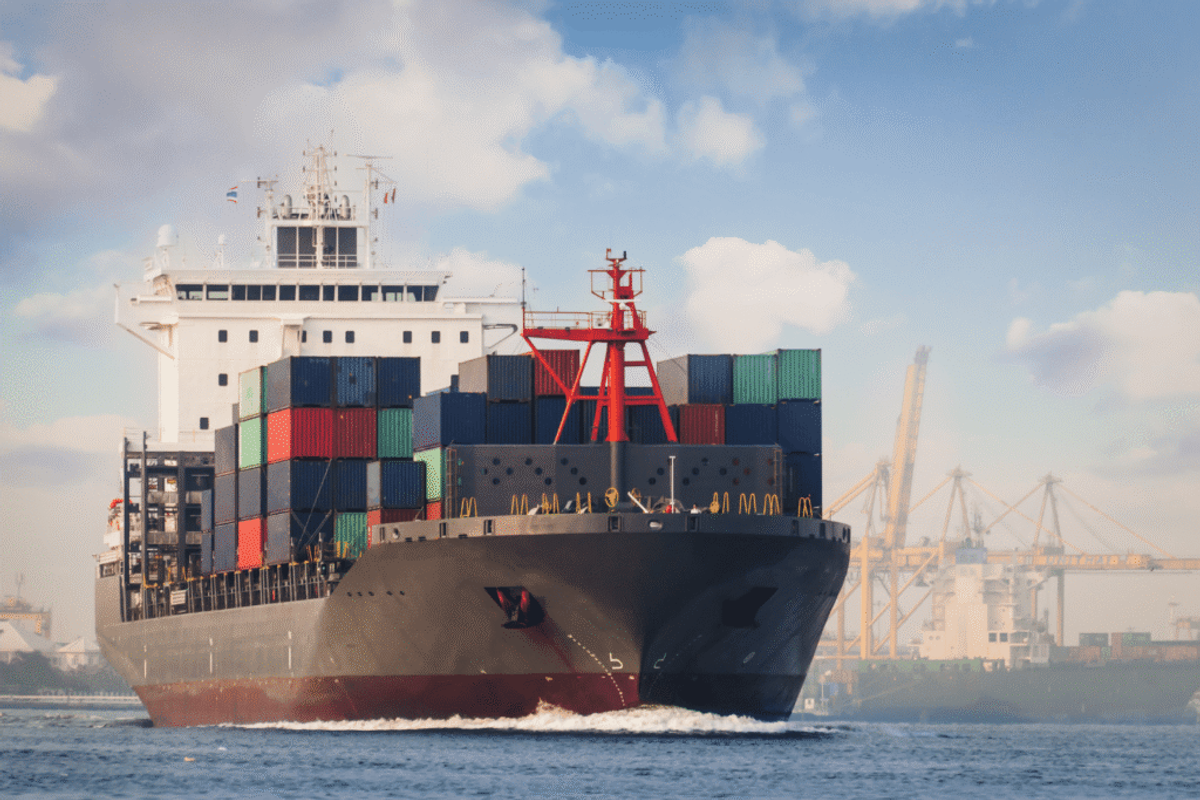 Large cargo ship loaded with colorful shipping containers sailing past cranes in a busy port
